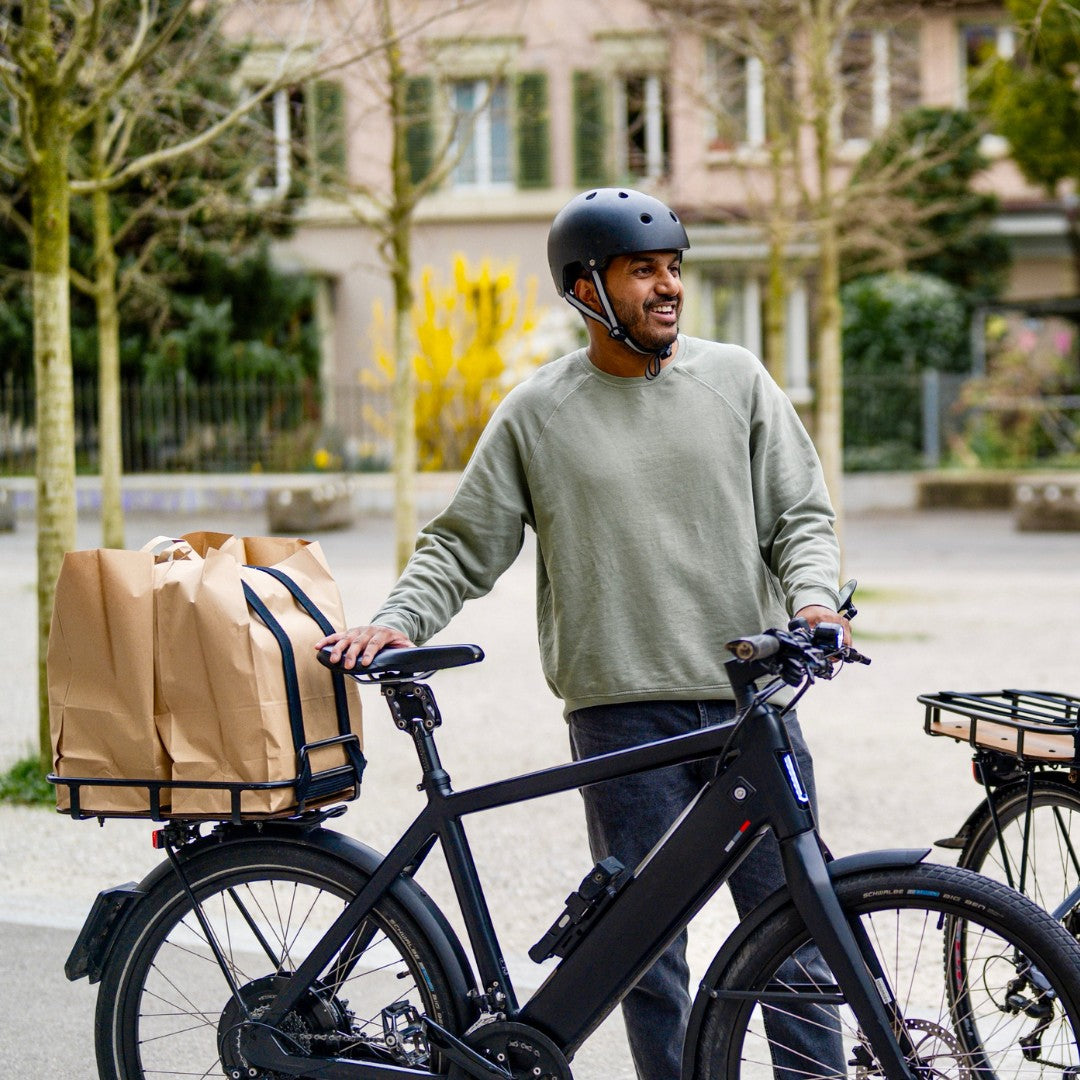 PACKI Velokorb auf E-Bike. Mann der lacht hält das Velo. Der Velokorb ist mit zwei Papiertaschen beladen. 